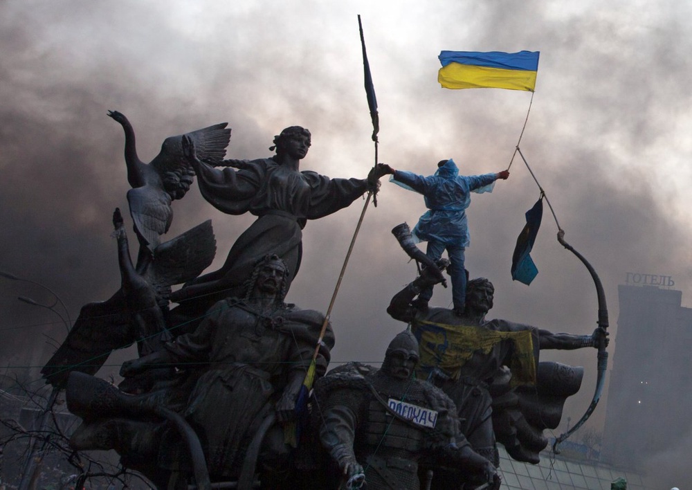 Антиправительственный протестующий держит государственный флаг Украины, взобравшись на памятник основателям Киева. ©REUTERS